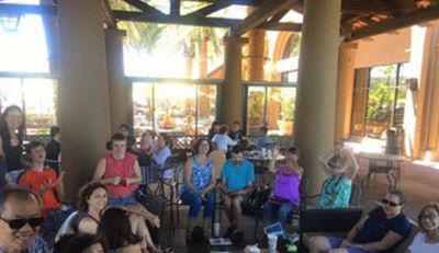 Group of people seated at an outdoor gathering, beneath a shaded area with pillars, enjoying the sunny day.