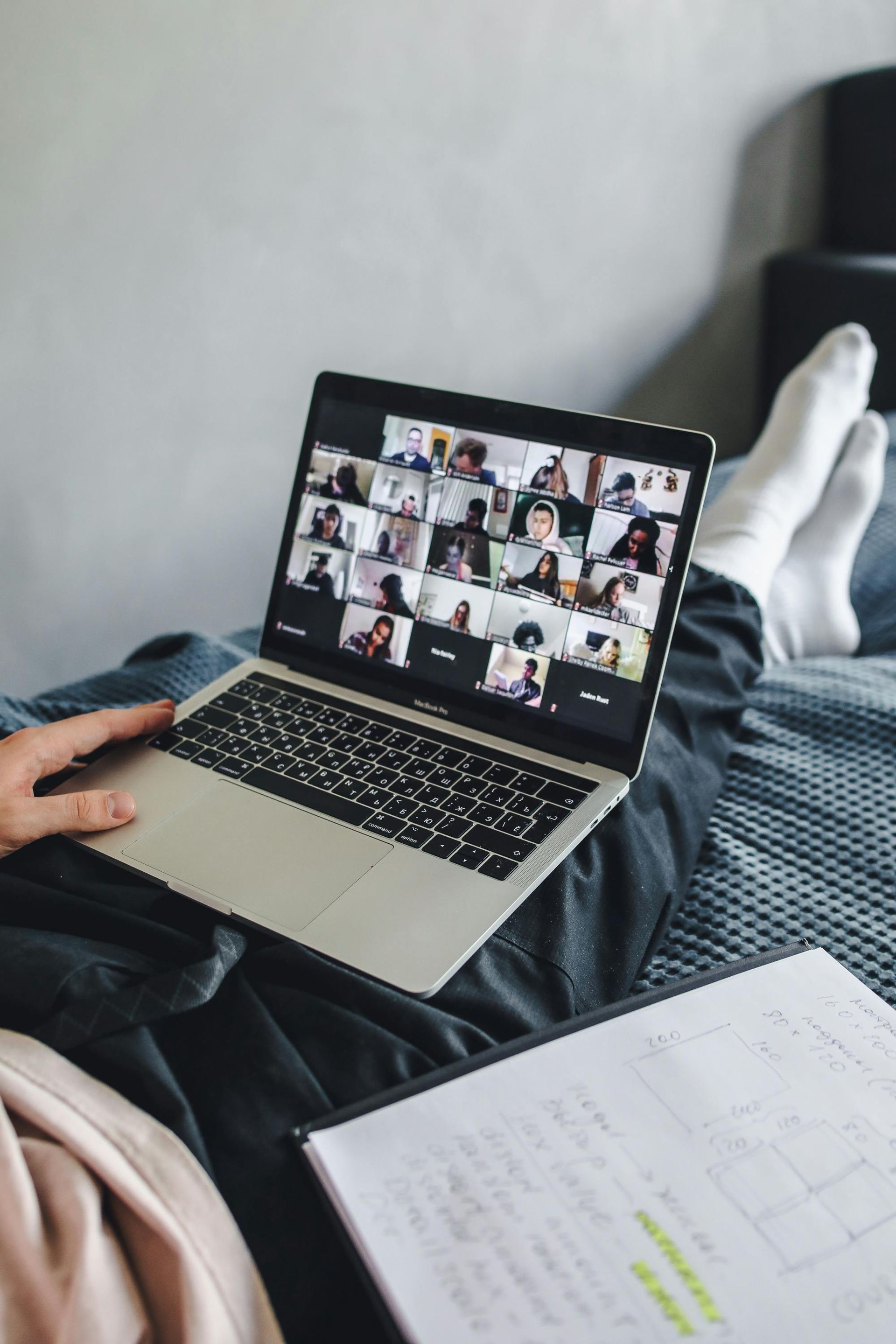 Person on bed, laptop showing video conference, notebook in lap.