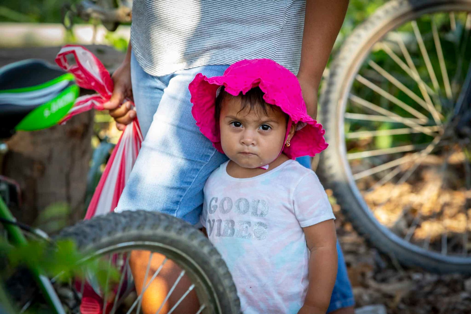 A toddler with a serious expression wears a bright pink sun hat and white shirt while standing in front of an adult.