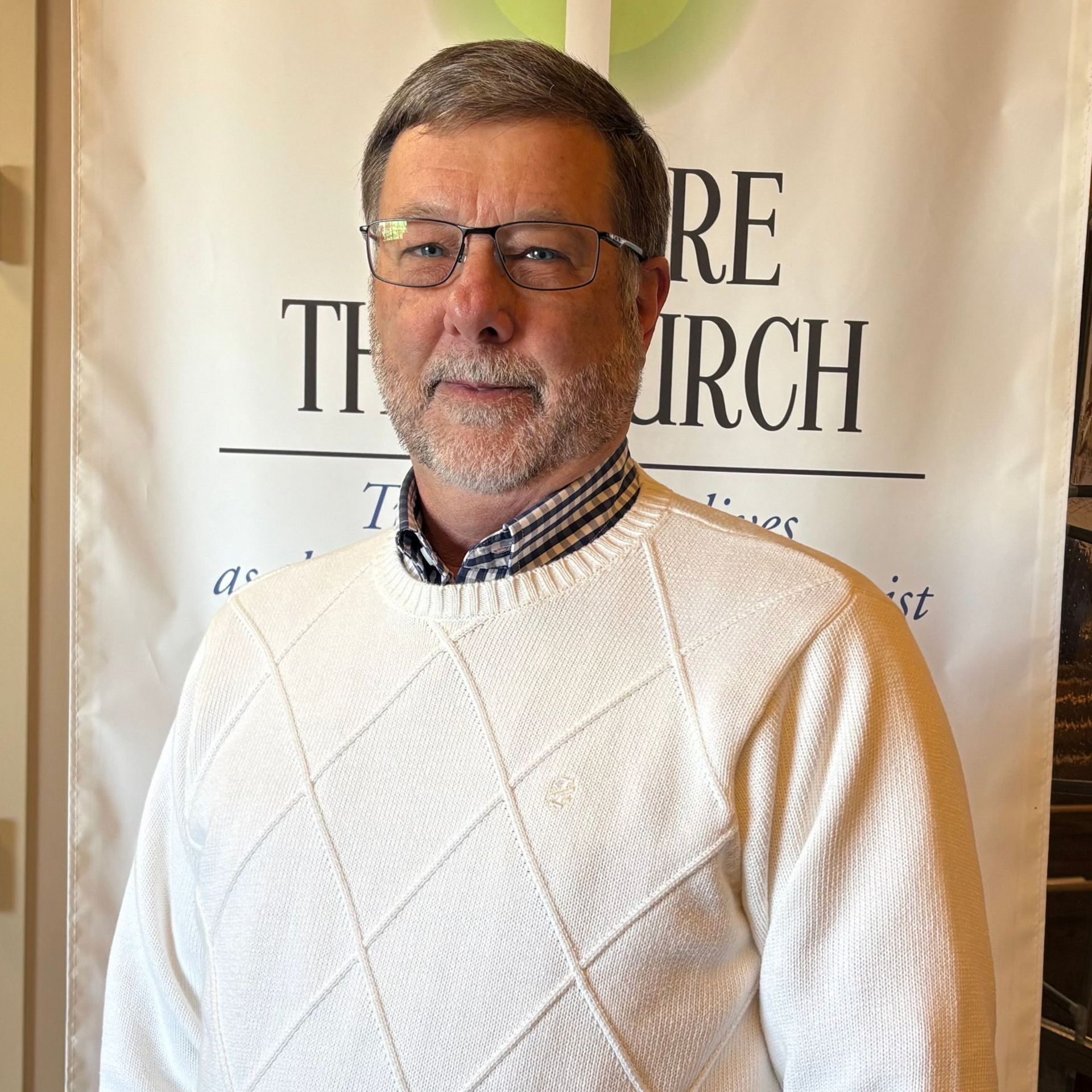 Man with glasses and white sweater, in front of a church banner.