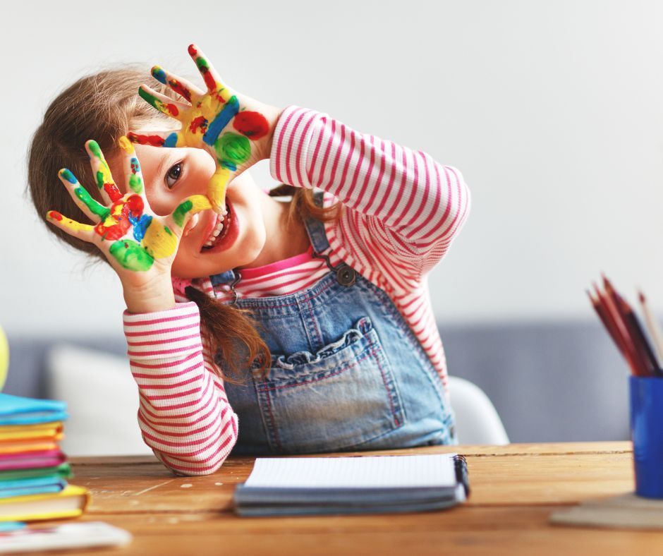 Child with painted hands making a playful face, sitting at a wooden table with books.