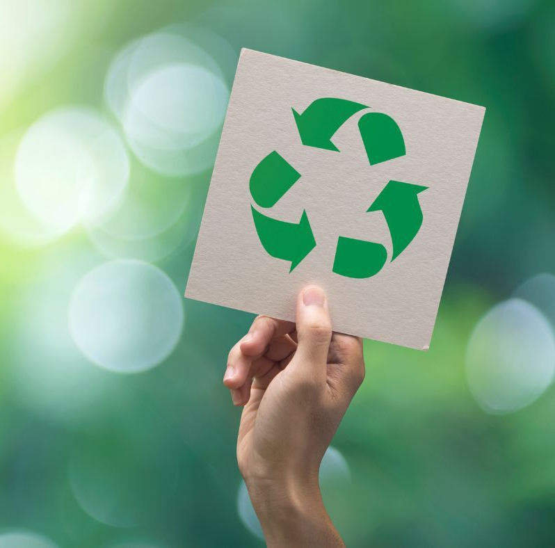 Hand holding a recycled paper sign with a green recycling symbol against a blurred green background.