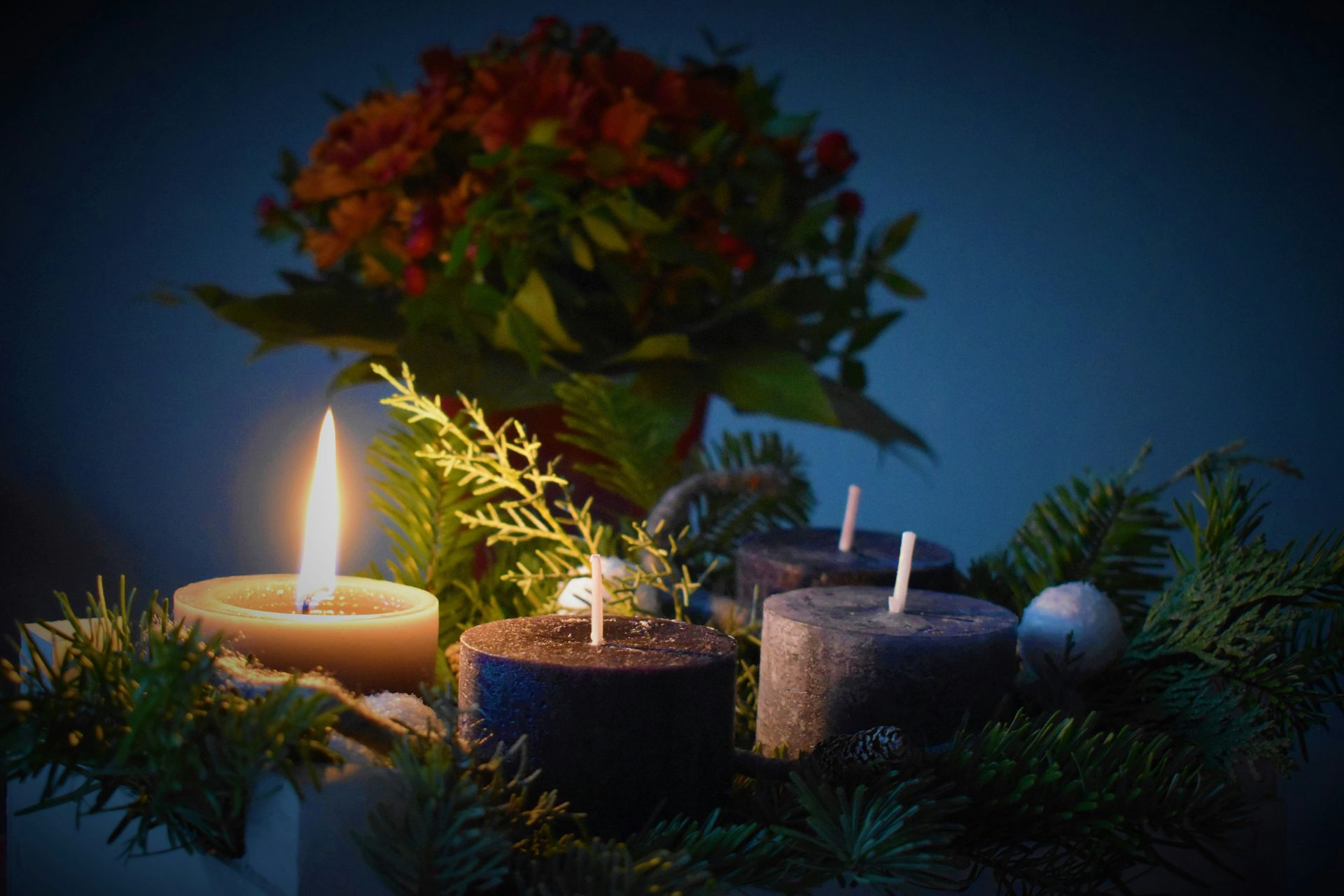 Advent wreath with one lit candle, other candles unlit, greenery, and flowers, blue background.