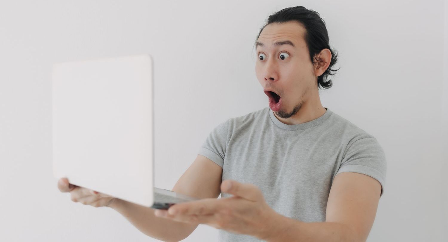 A man is holding a laptop computer in his hands and making a surprised face.