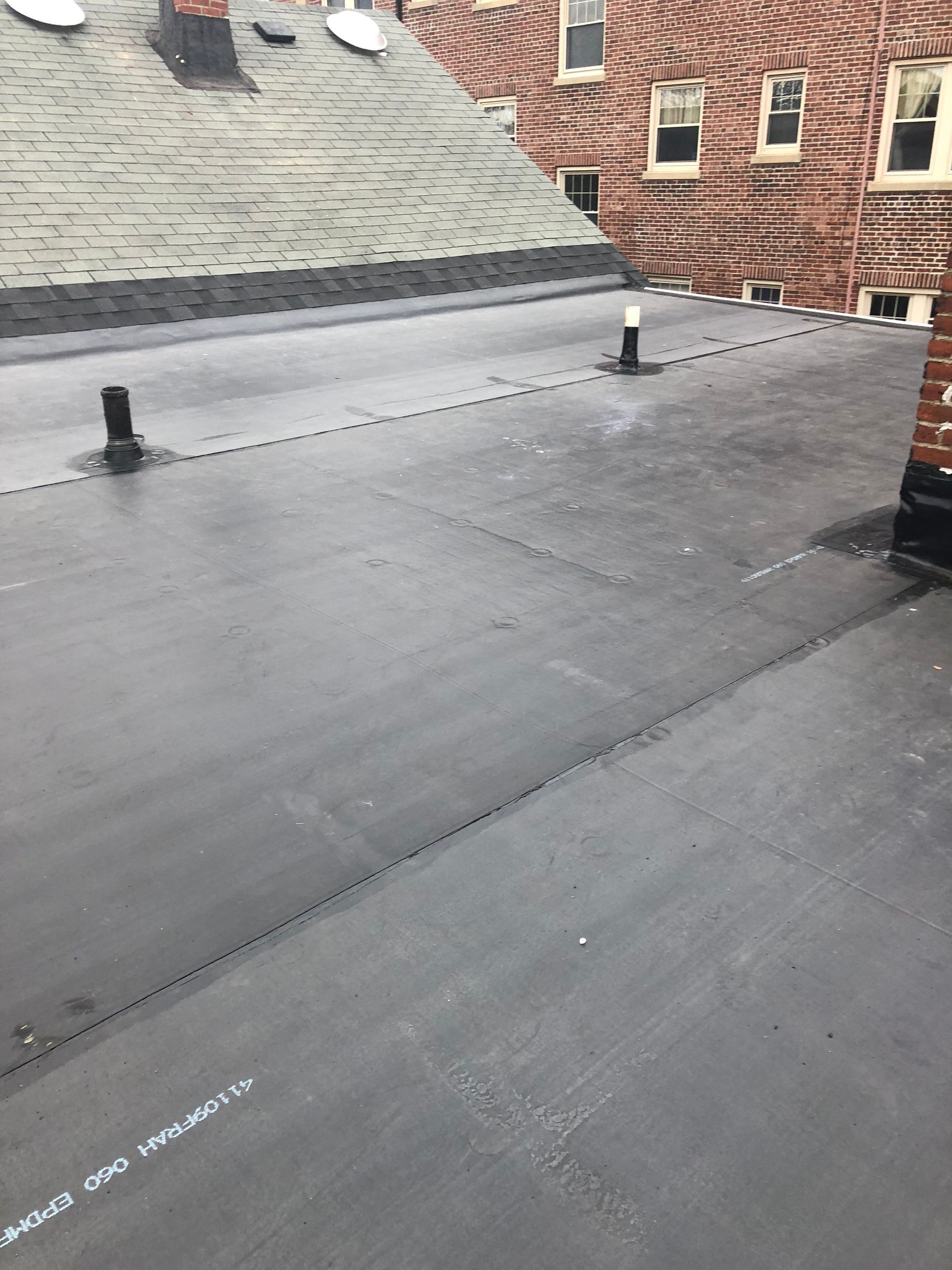 Flat rooftop with black membrane, vents, and adjacent brick buildings under an overcast sky
