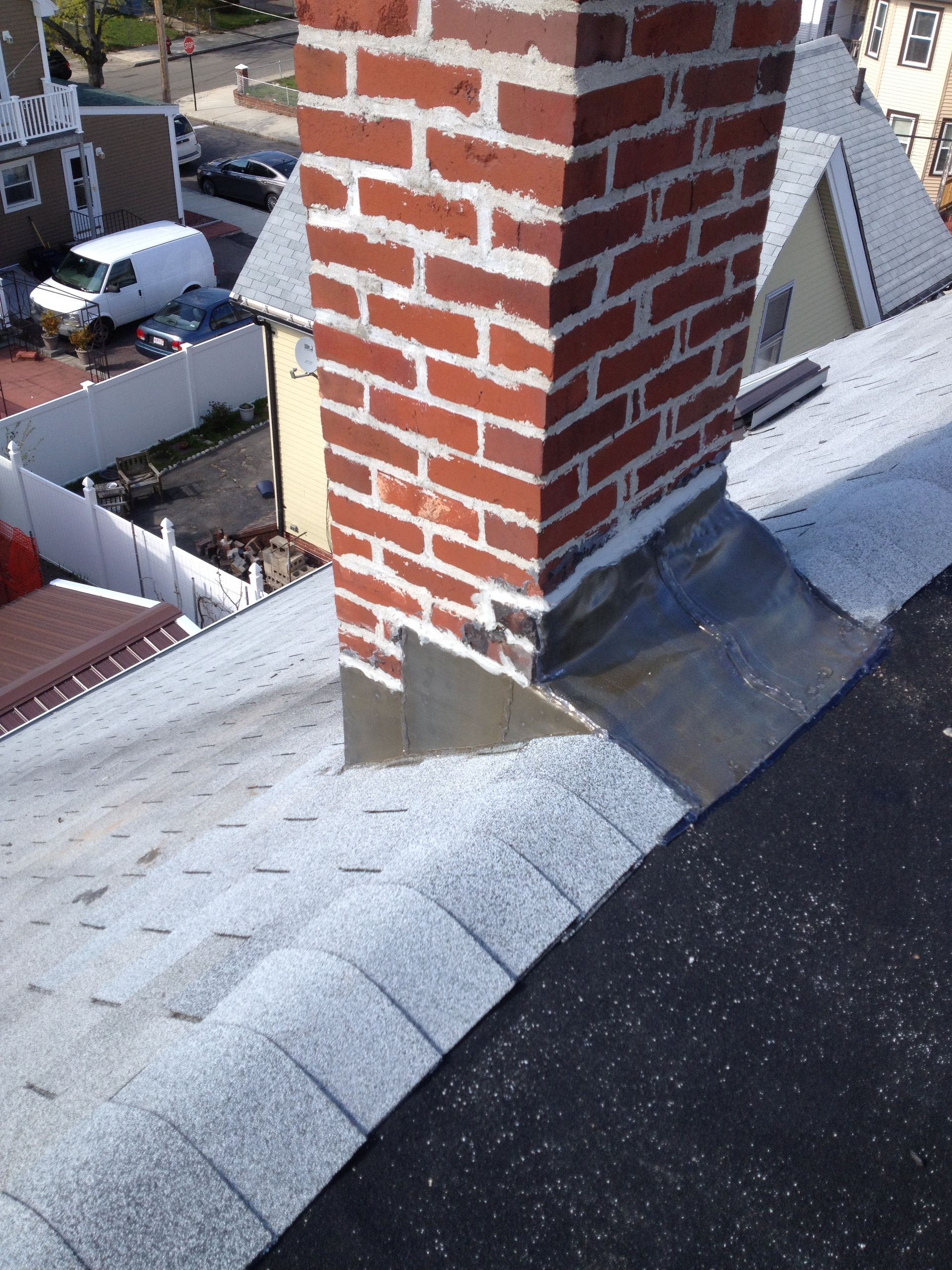 Red brick chimney rising through a gray shingled rooftop with nearby houses visible below