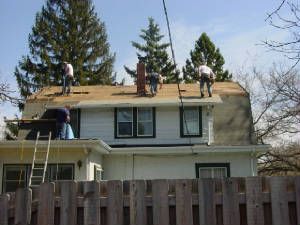 Workers repairing a roof on a two-story house behind a wooden fence