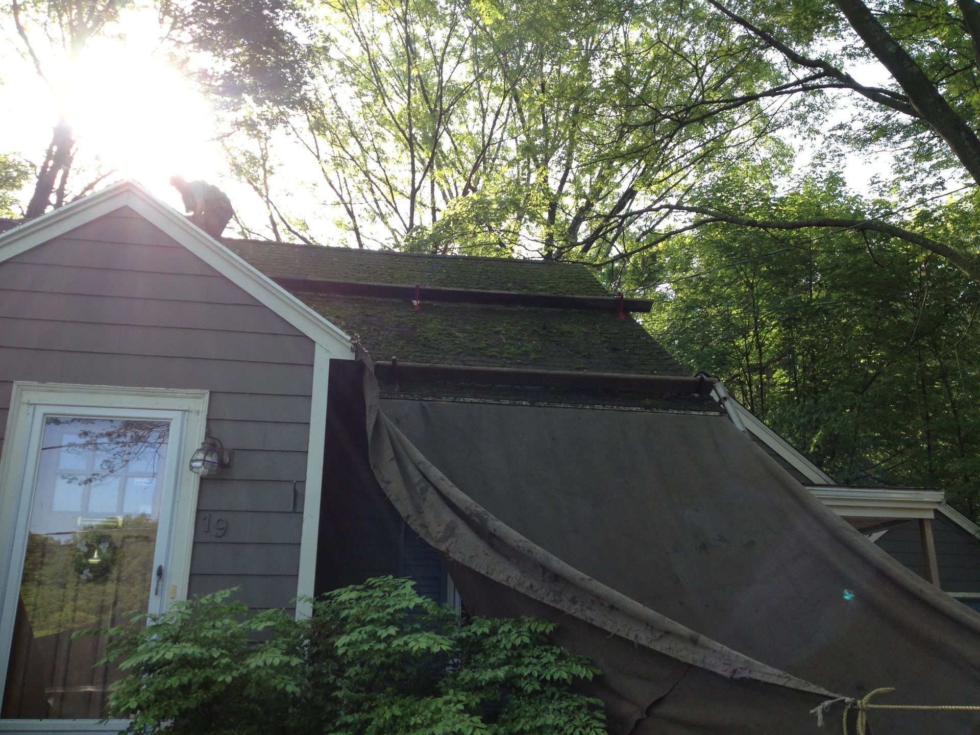 Damaged house roof with a large torn tarp draped over it, surrounded by trees and sunlight.