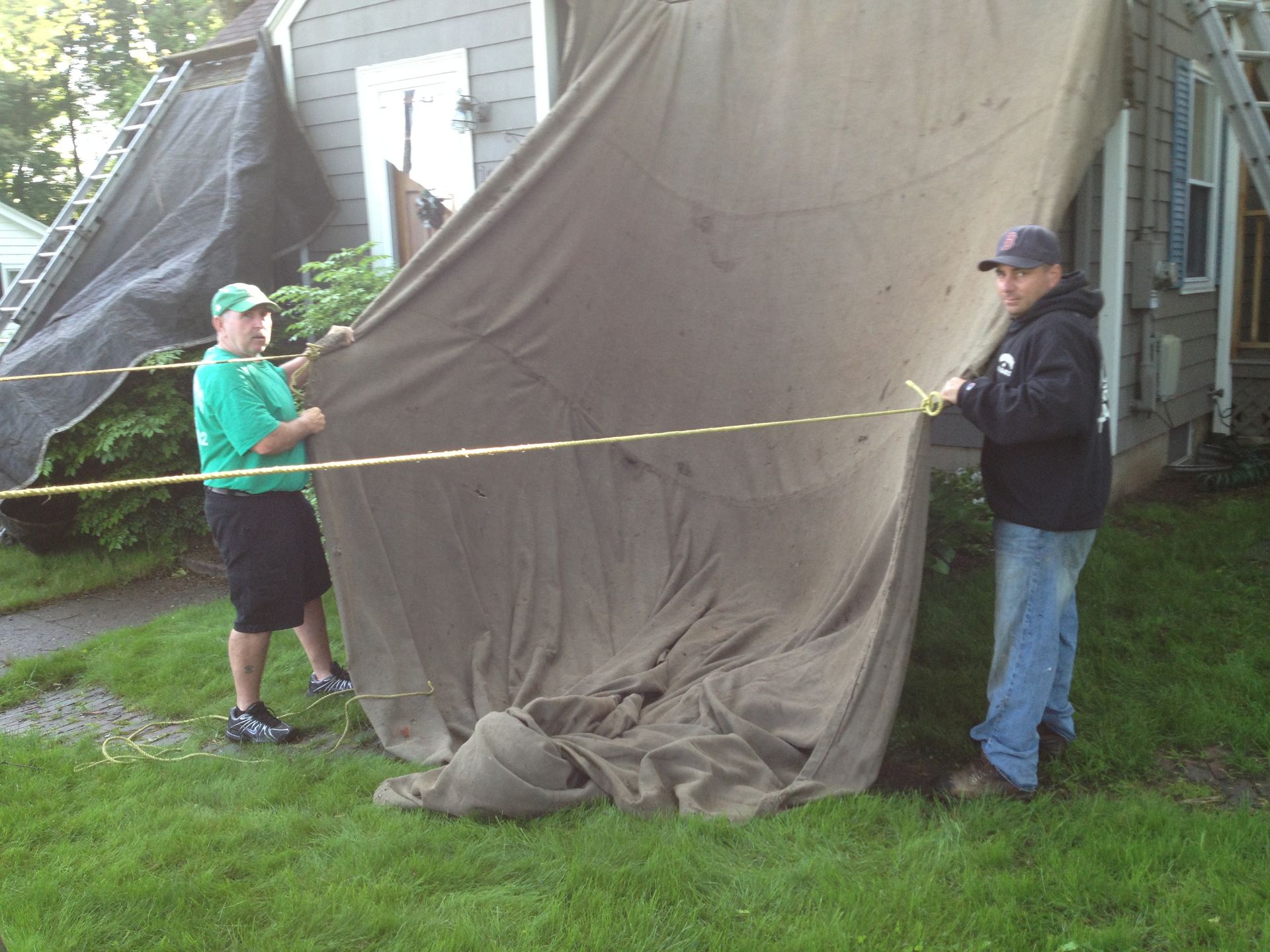 Two people stretching a large brown tarp beside a house, one holding a yellow measuring tape.