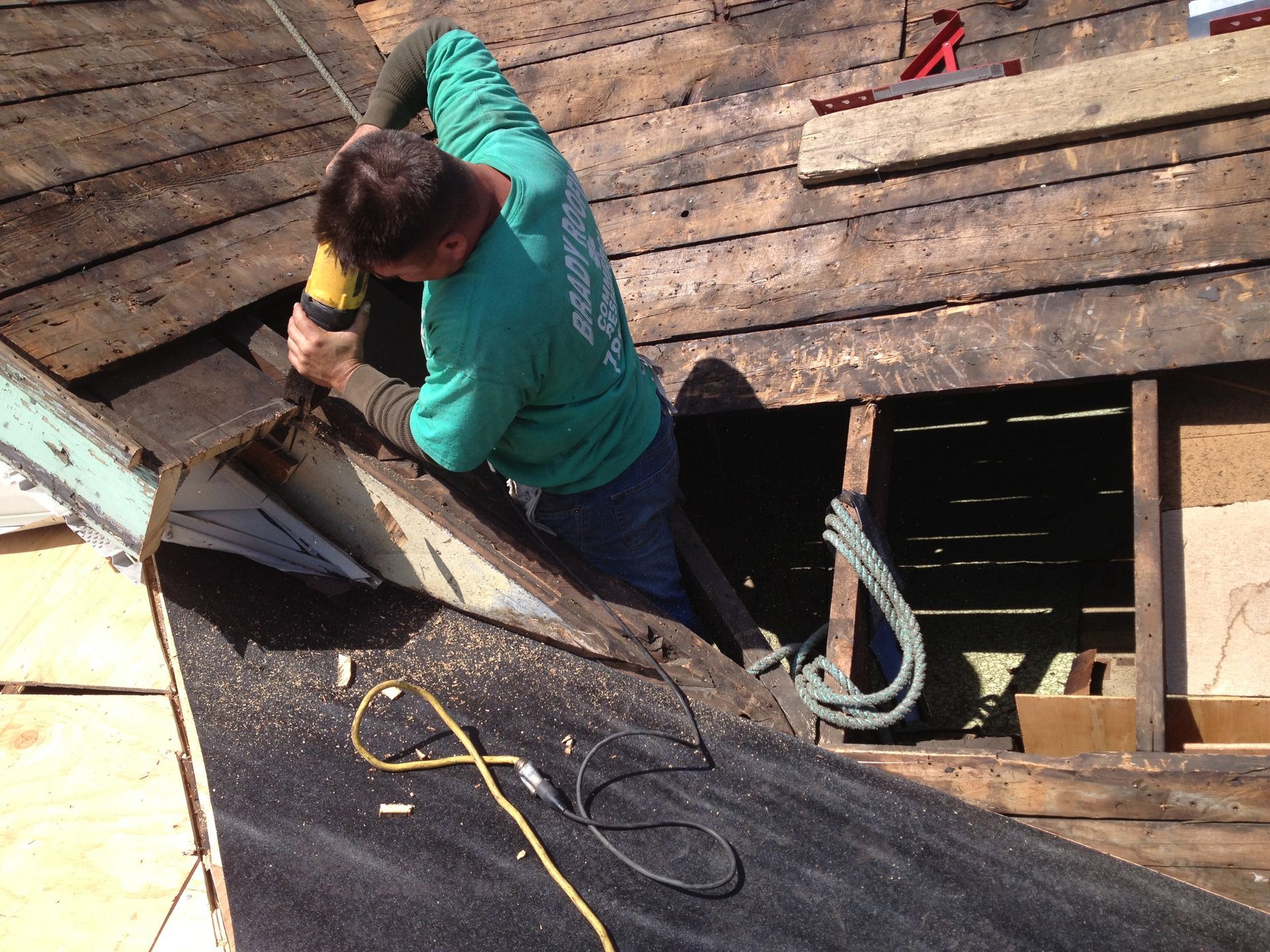 Person using a tool to repair a weathered roof, leaning over a damaged section with cords nearby