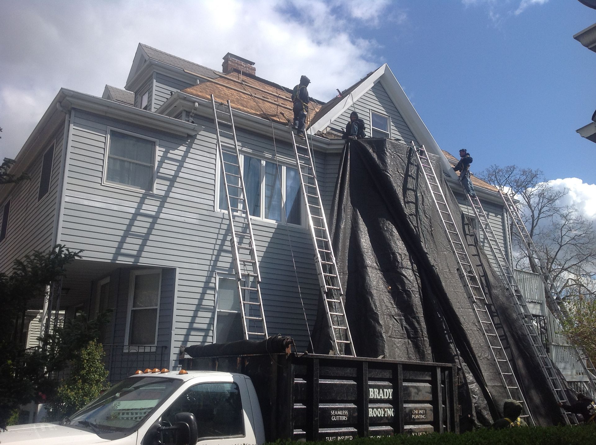 Laddered house with roof repair, large dumpster, and a work truck in front under a blue sky.