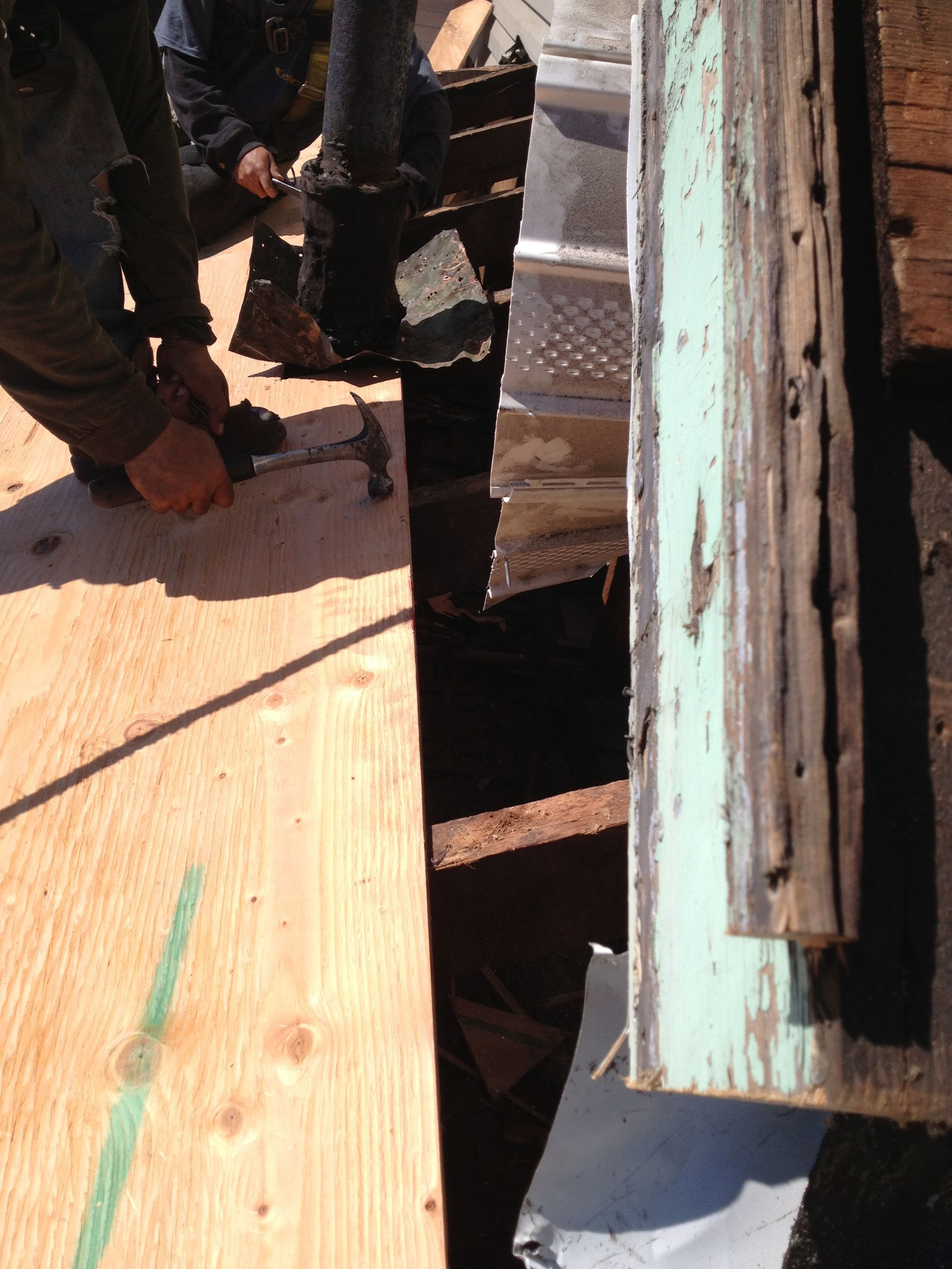 Worker kneeling on a wooden roof, handling boards beside a narrow gap and weathered siding.
