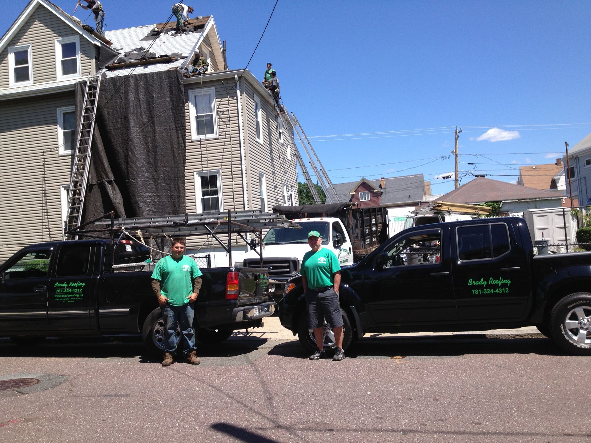Two workers beside black pickup trucks on a street, with a damaged house and debris in the background