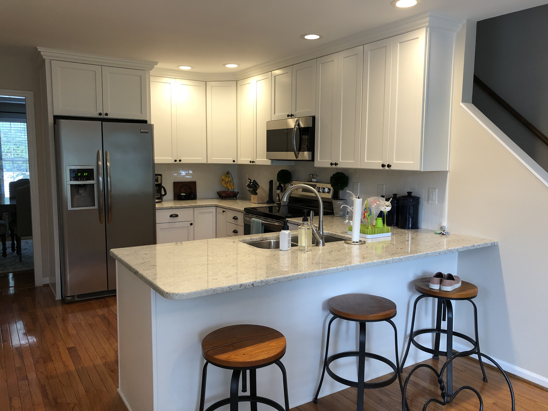 A kitchen with white cabinets and stools and a stainless steel refrigerator.