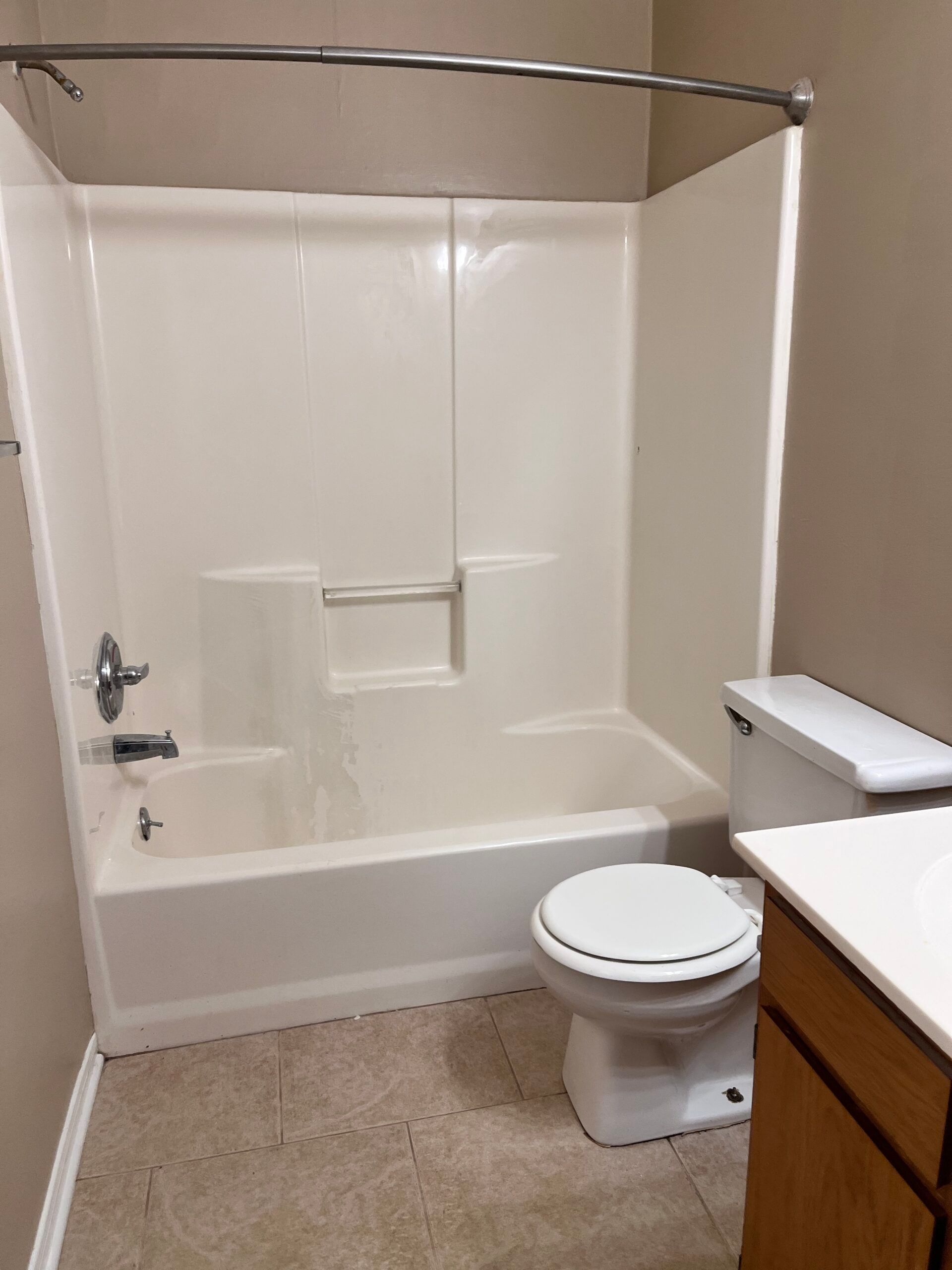 Bathroom with white tub/shower combo, toilet, beige walls and floor, and wooden cabinet.