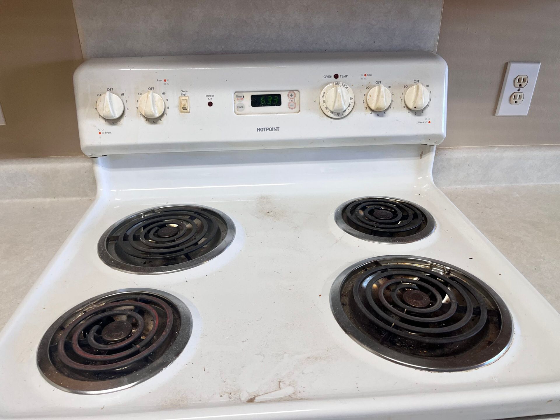 White electric stove with four burners and knobs, a digital clock, and a dirty surface.