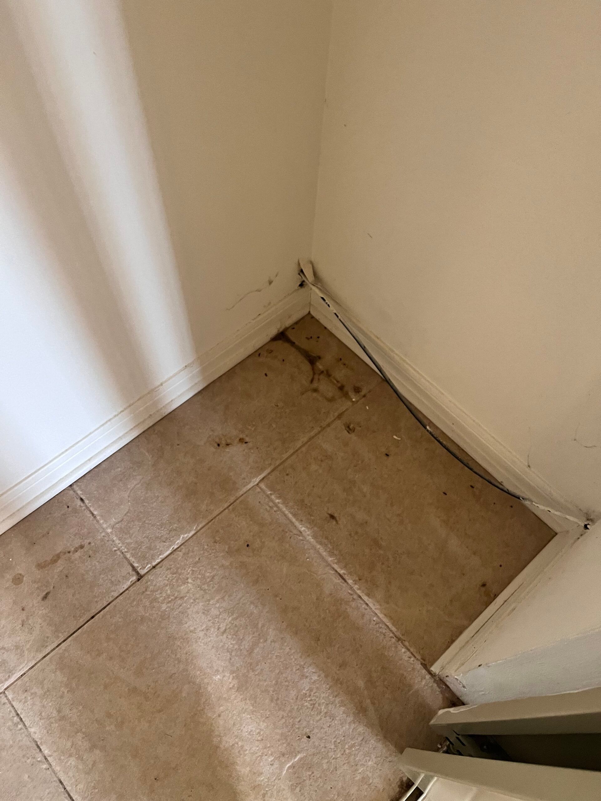 Corner of a room with tan tile floor and white walls, showing debris and potential water damage.