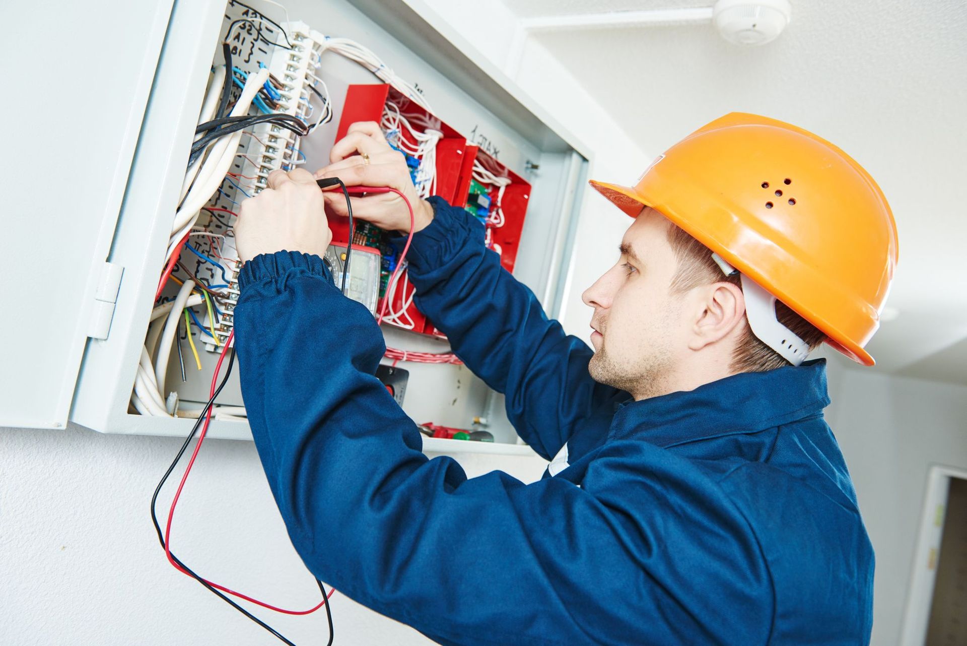 Electrician in orange hard hat wiring an open electrical panel with red and black wires