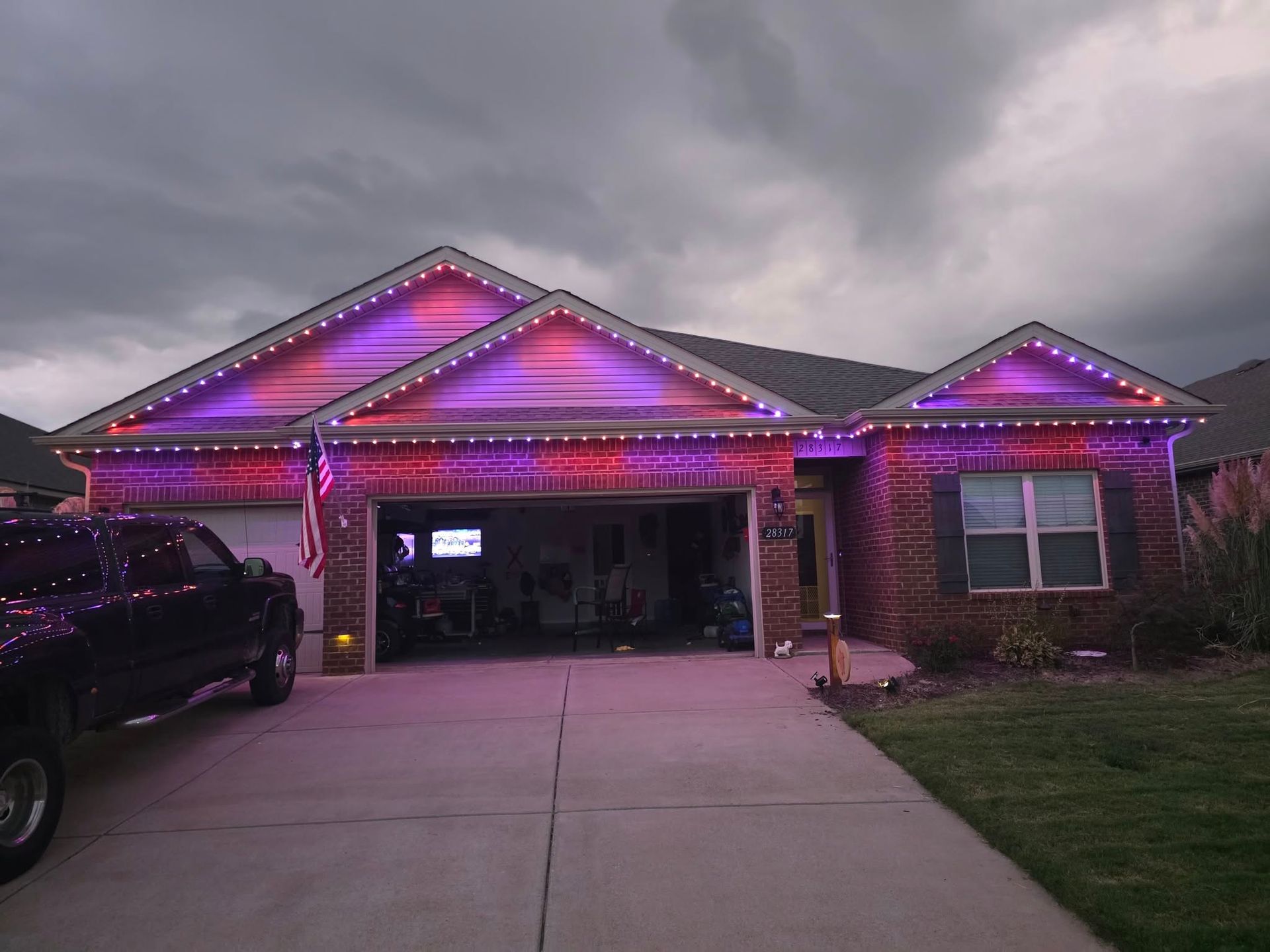 House with red and purple lights on roof and along the eaves, with a cloudy sky. A truck is parked in the driveway.