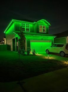 Two-story house illuminated with green lights, including roofline, garage, and ground. White car parked in driveway. Night setting.