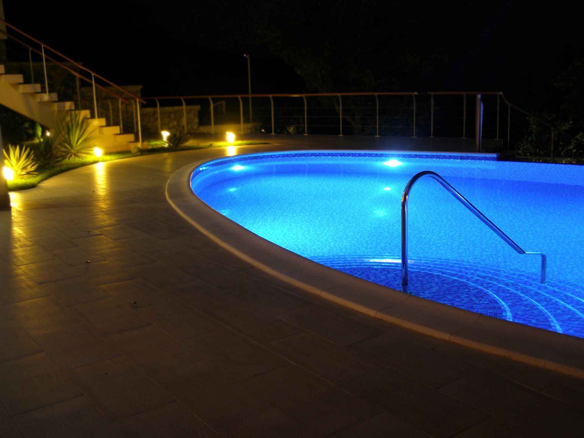 Night view of a curved pool with blue underwater lights, lit pathway, and stairs.