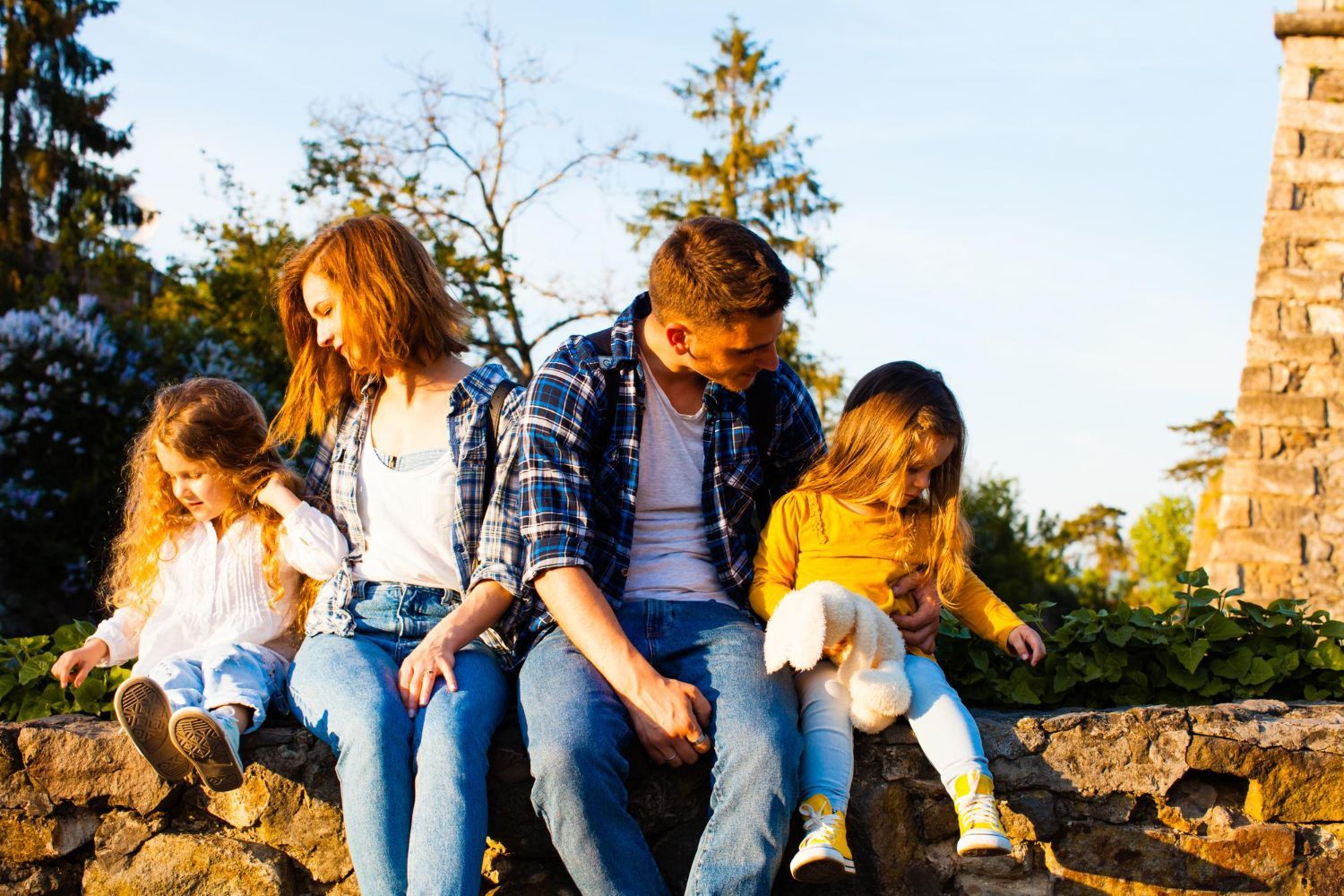 Four friends sit on a stone wall outdoors in warm sunlight, chatting and looking down.