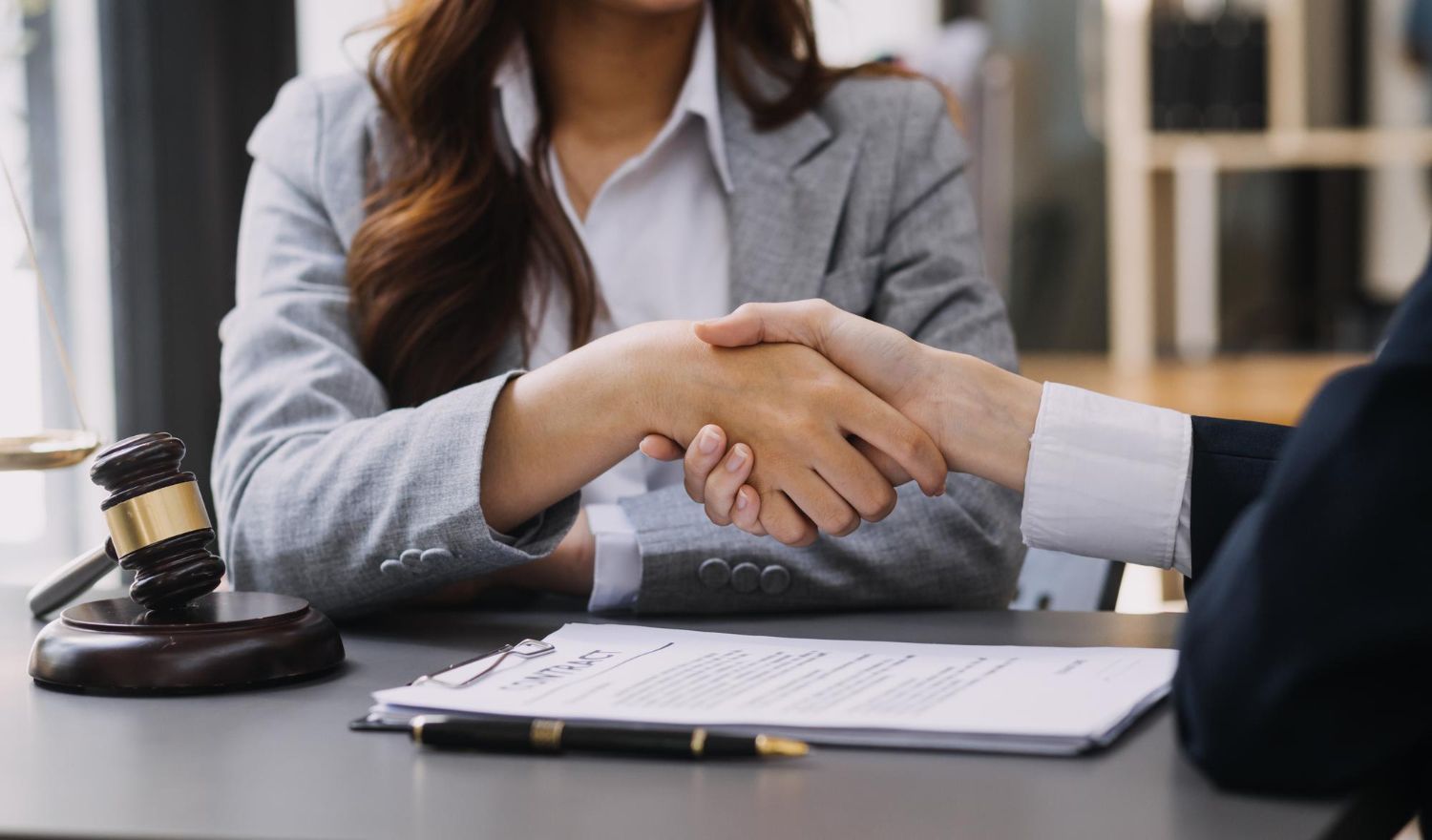 Handshake between two people over a desk with a gavel and documents, suggesting a legal agreement.