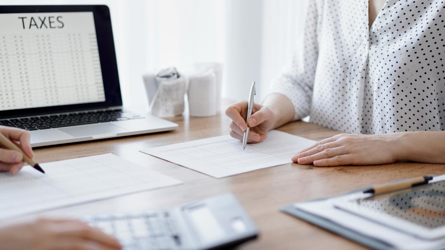 Person writing at a desk beside a laptop showing “TAXES” and paperwork