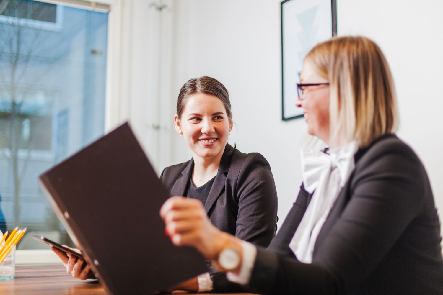 Two coworkers in a bright office discussing a black folder across a desk