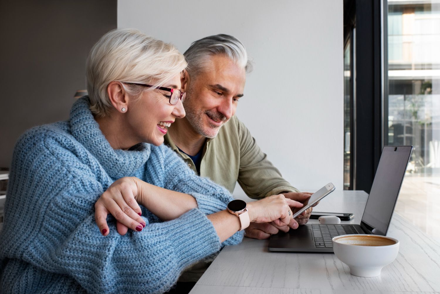 Two people smiling at a laptop and tablet at a cafe table by a window