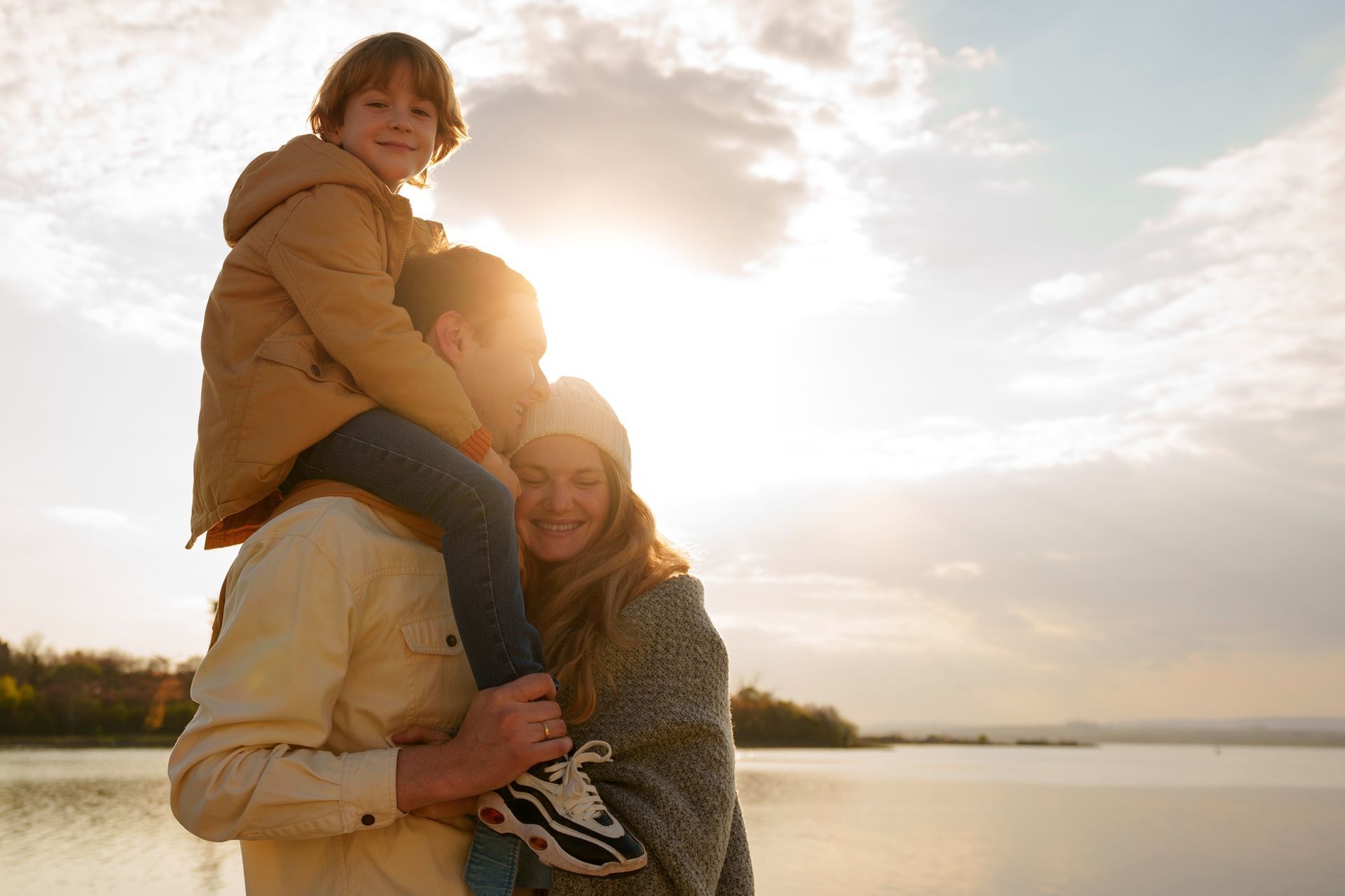 Family embracing by a lakeshore at sunset, with one child on an adult’s shoulders.