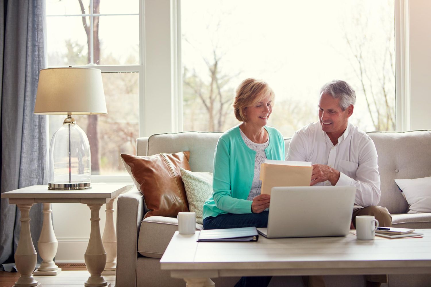 Two people reviewing documents on a couch beside a laptop in a bright living room