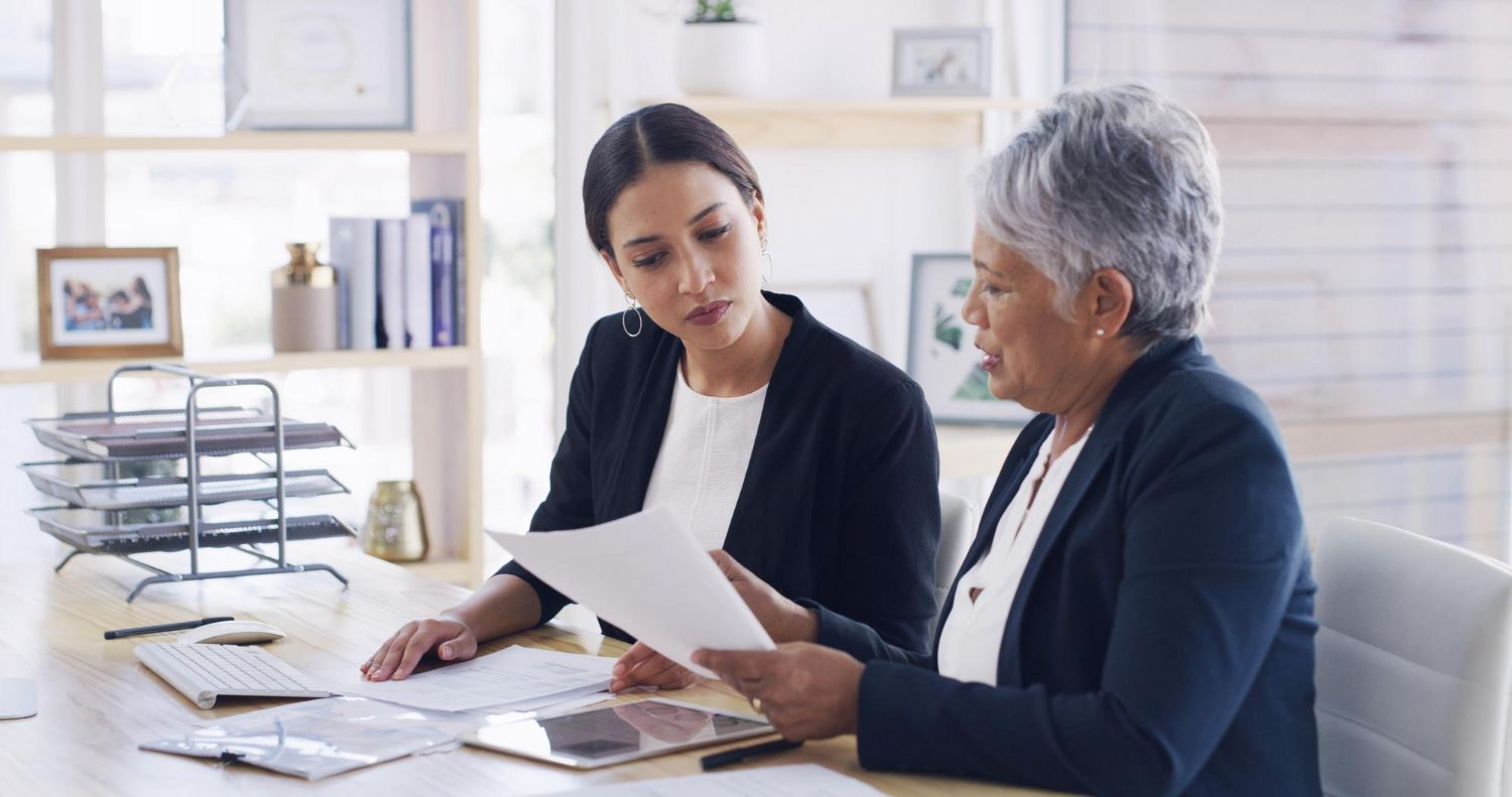 Two women review papers at a desk in a bright office with a file organizer and shelves behind them.