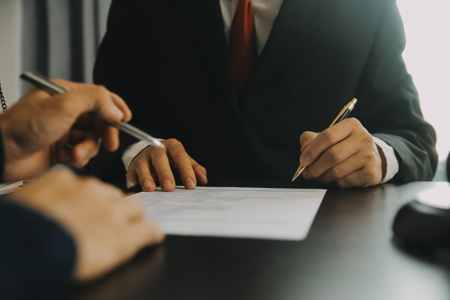 People in suits signing a document at a table, with hands and pens visible.