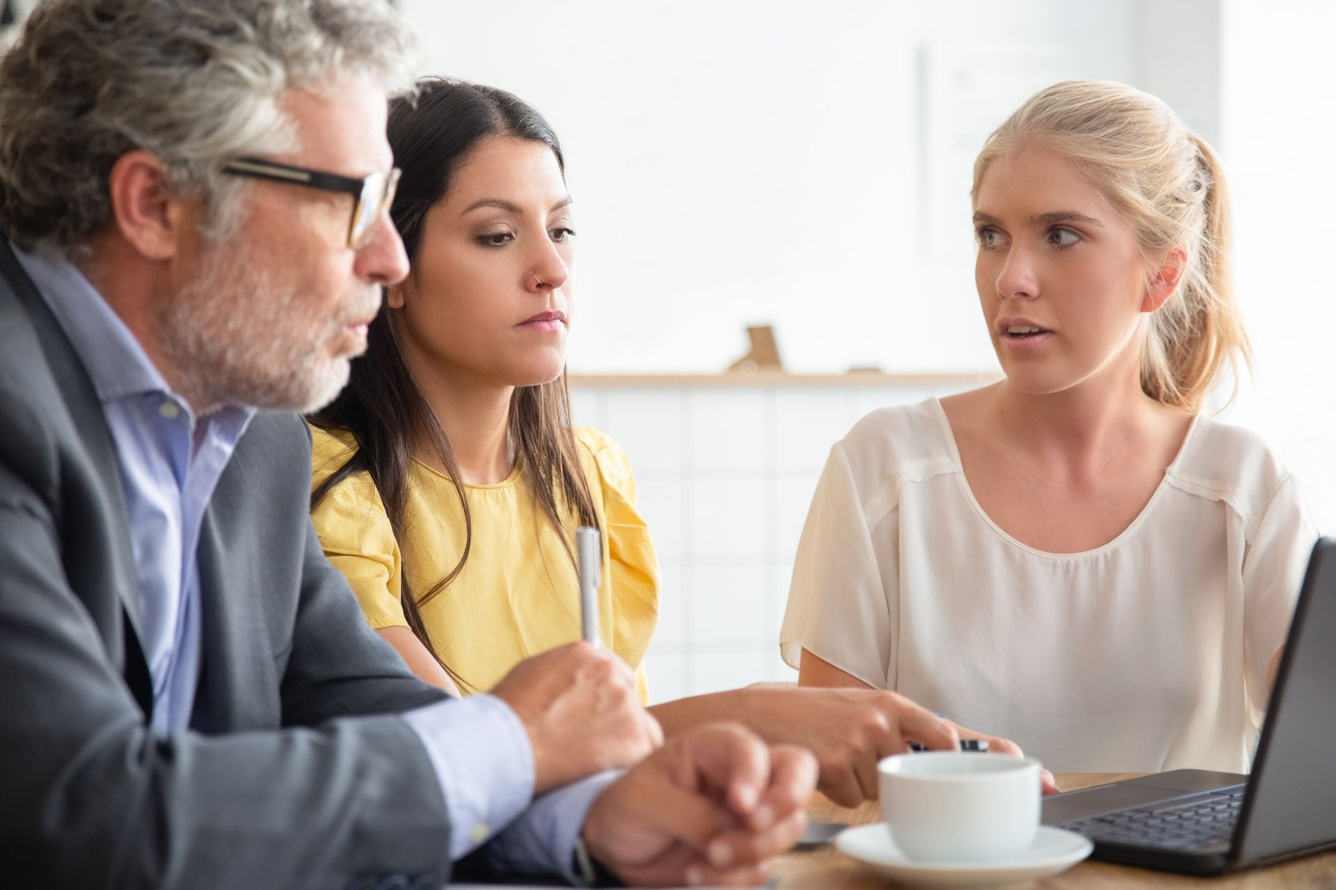 Three coworkers in a meeting, discussing beside a laptop and coffee cup in a bright office.