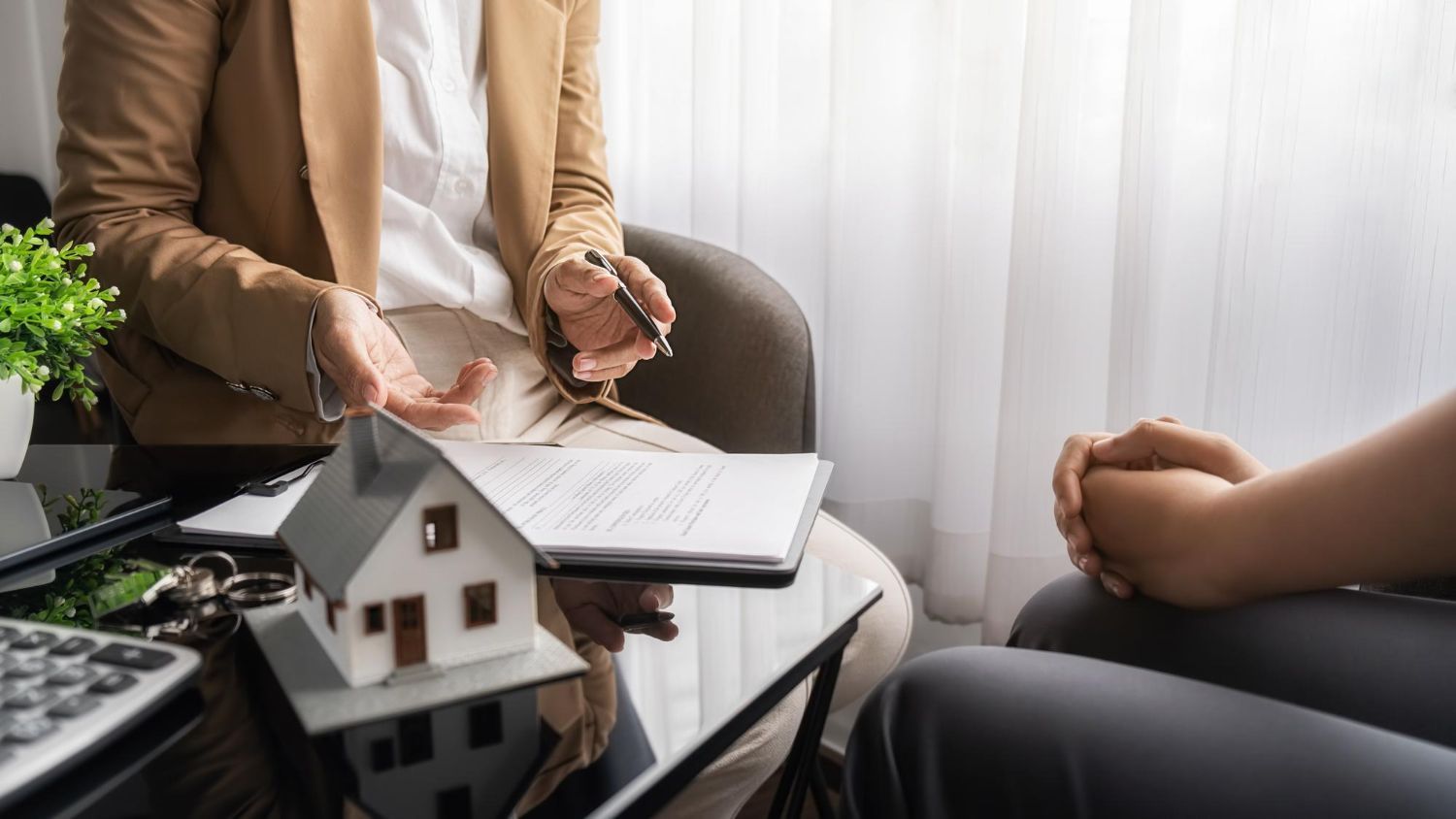 Two people in a meeting discussing a house model and papers at a table by a window