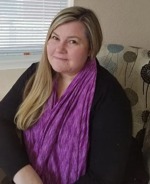 Woman seated indoors, wearing a black top and bright purple scarf, smiling softly near a window.