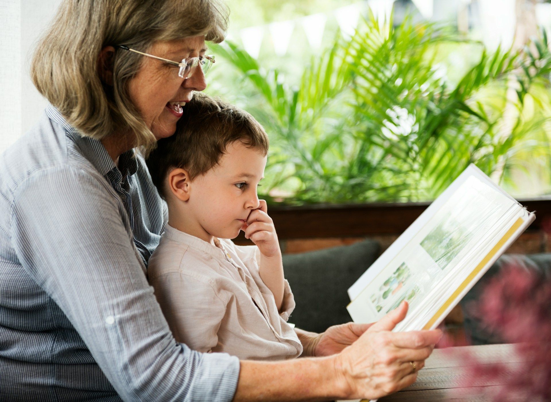 Older woman and young child reading a book together on a couch with greenery in the background
