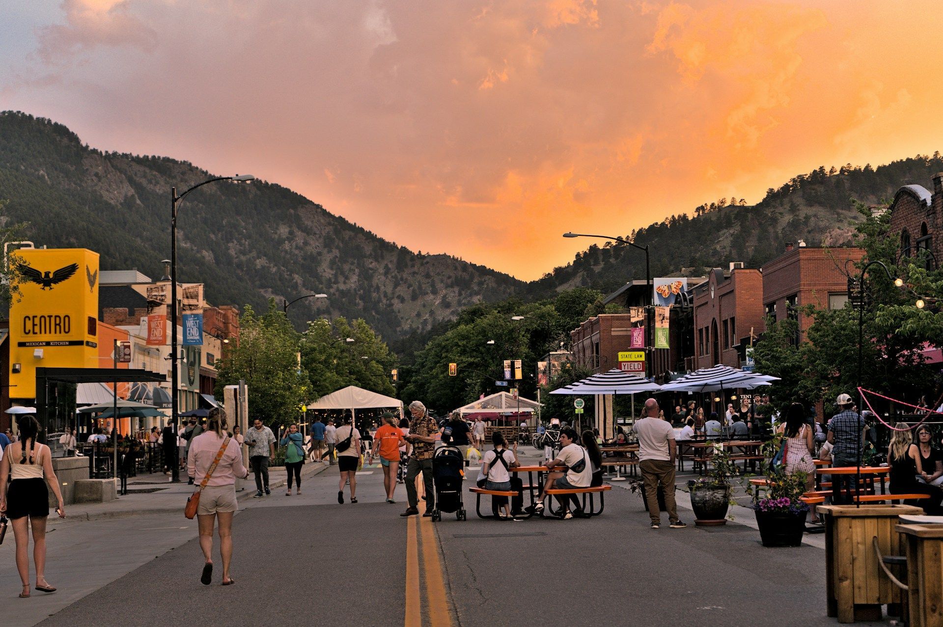 Busy mountain town street at sunset with outdoor diners and shops under an orange sky