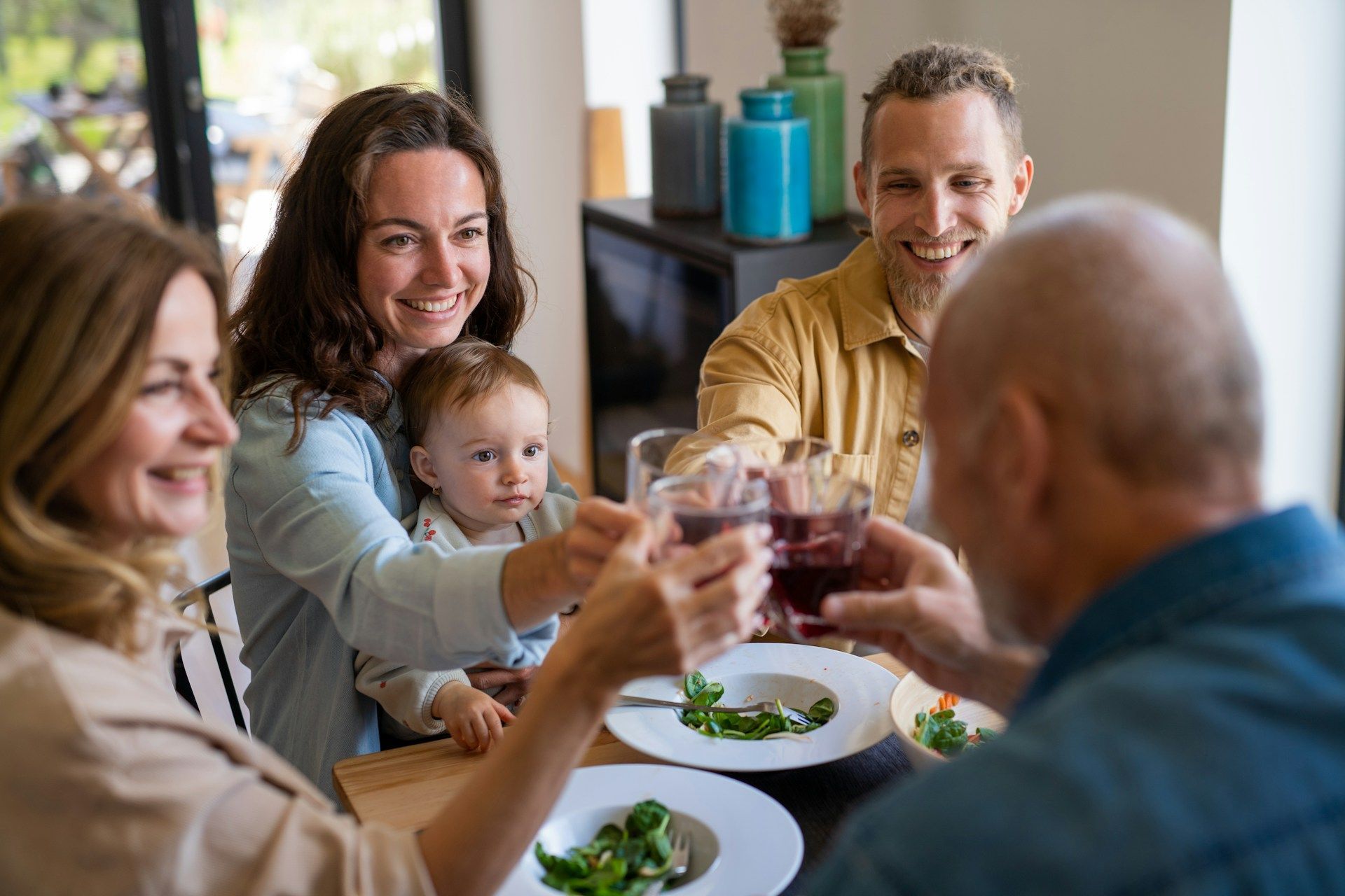 Family toasting at a dining table with a baby, plates of food, and drinks in a bright home dining room