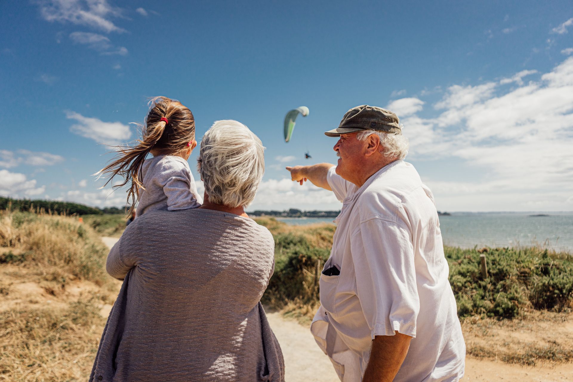 Three people on a coastal path, one pointing toward the sea under a bright blue sky.