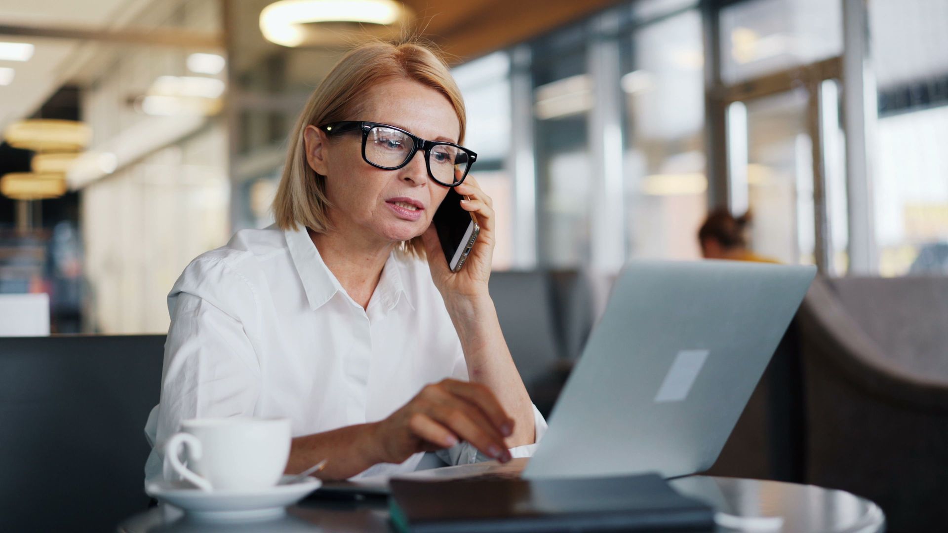 Woman in glasses working on a laptop and talking on a phone at a café table