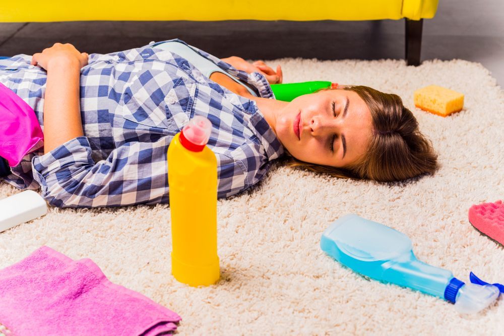 A person lies on a carpet surrounded by cleaning supplies, including a yellow bottle, blue spray, and cloths.