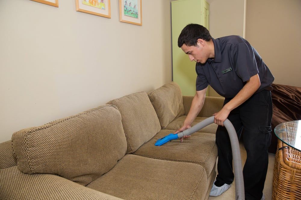 A man is cleaning a couch with a vacuum cleaner.