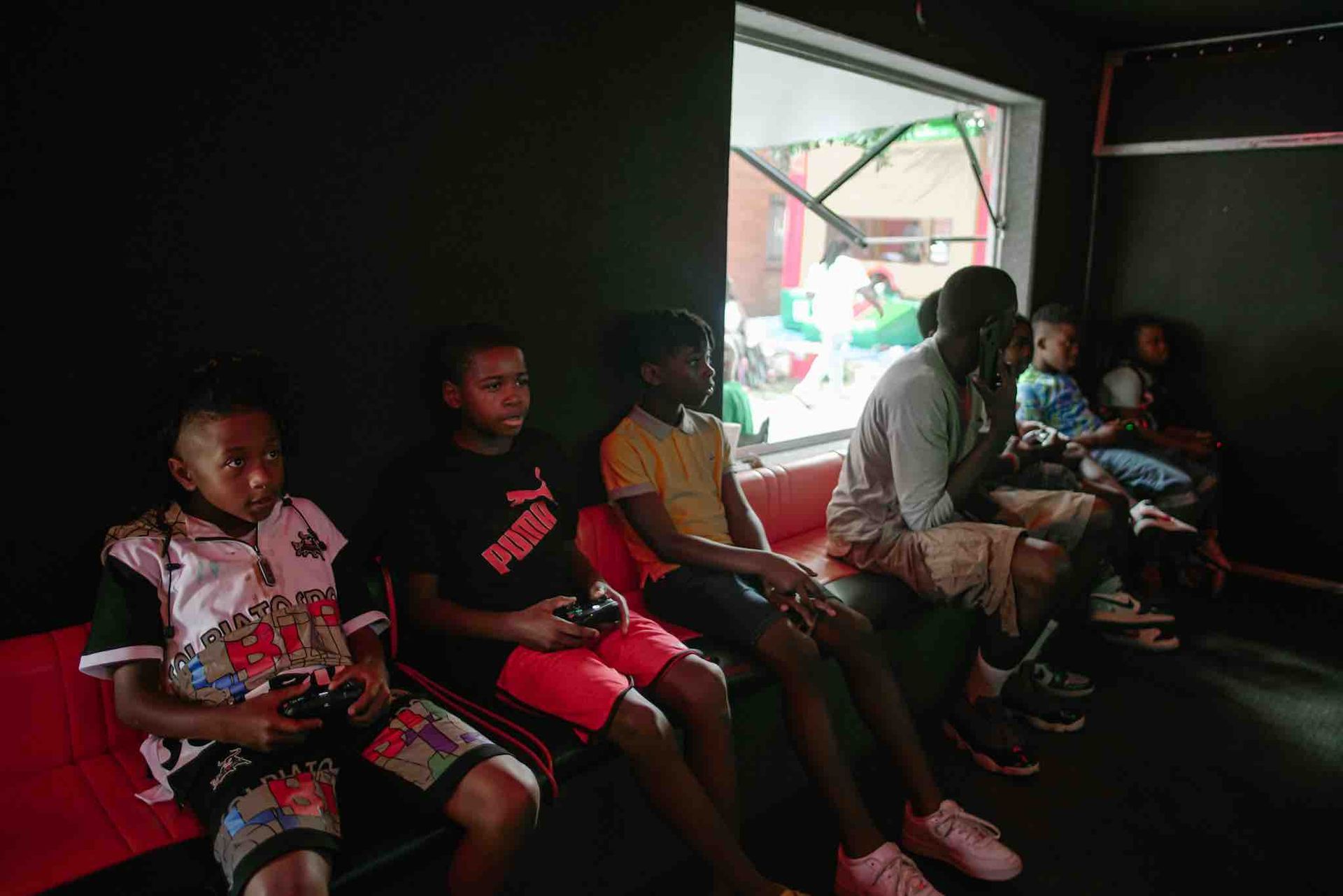 Several people sitting and holding game controllers in a dark room with a window showing a colorful outdoor area.