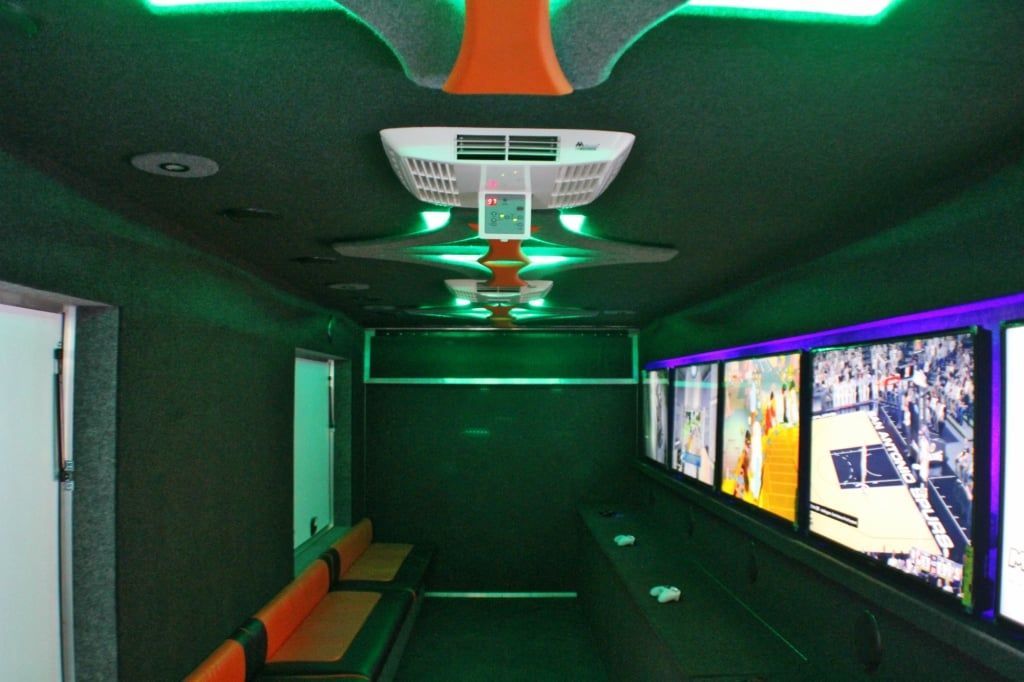 Interior of a gaming trailer, featuring several monitors, seating, and green and orange lighting.