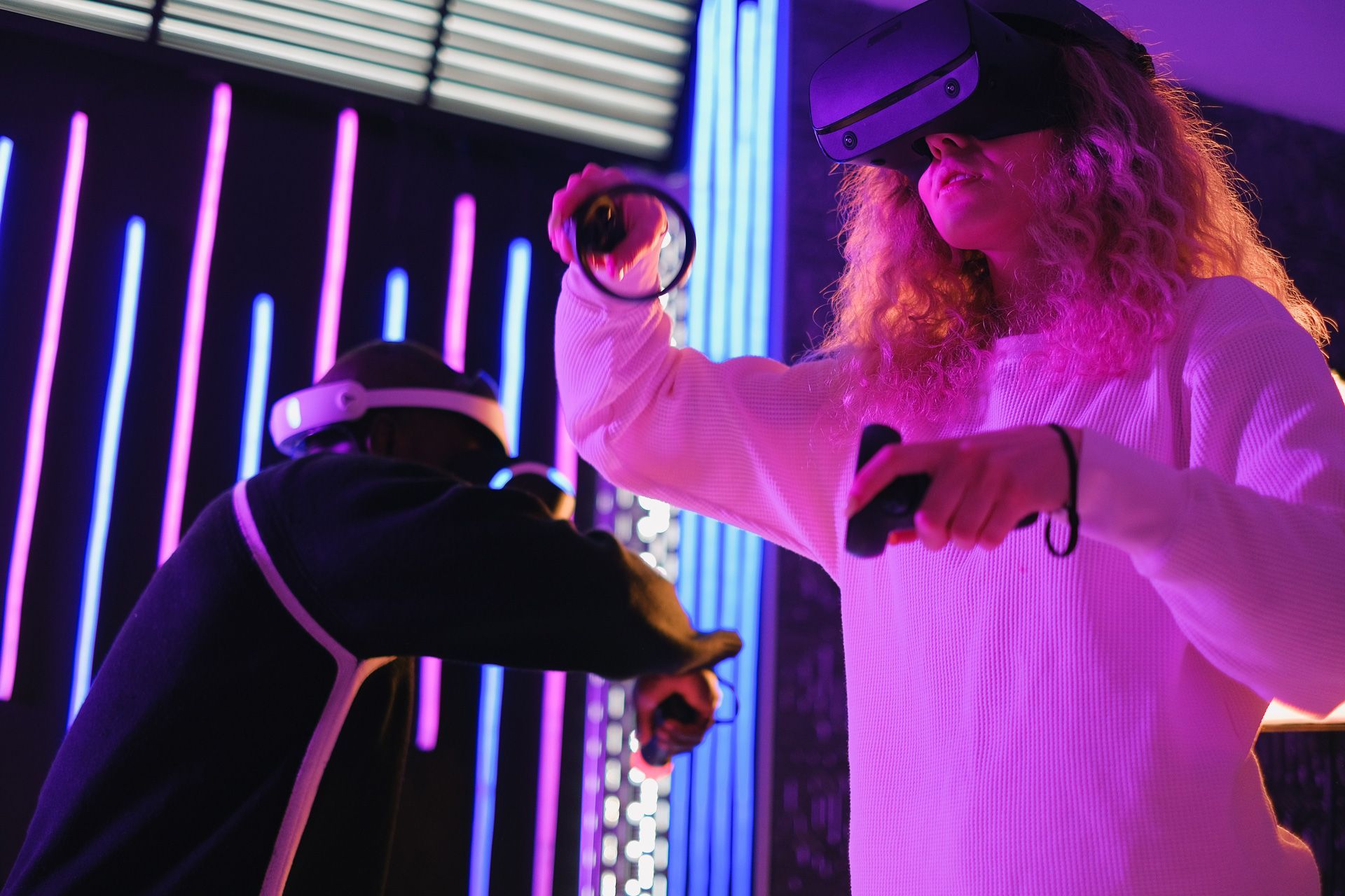 Two people wearing VR headsets and using controllers in a neon-lit room, possibly playing a game.