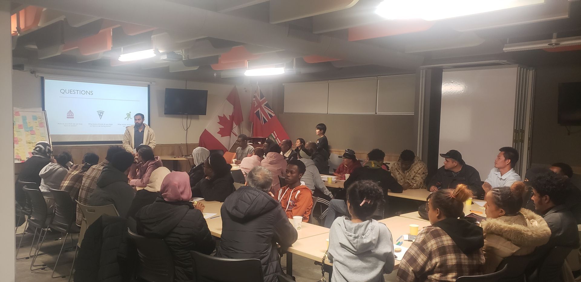 People seated at tables in a room, watching a presentation on a screen. A Canadian flag is visible.