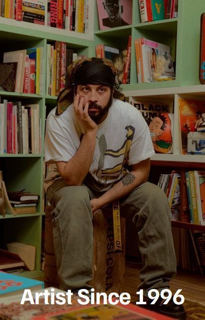 Man with beard, black headscarf, white shirt, sitting on a wooden box, leaning on hand. Bookshelves in background.
