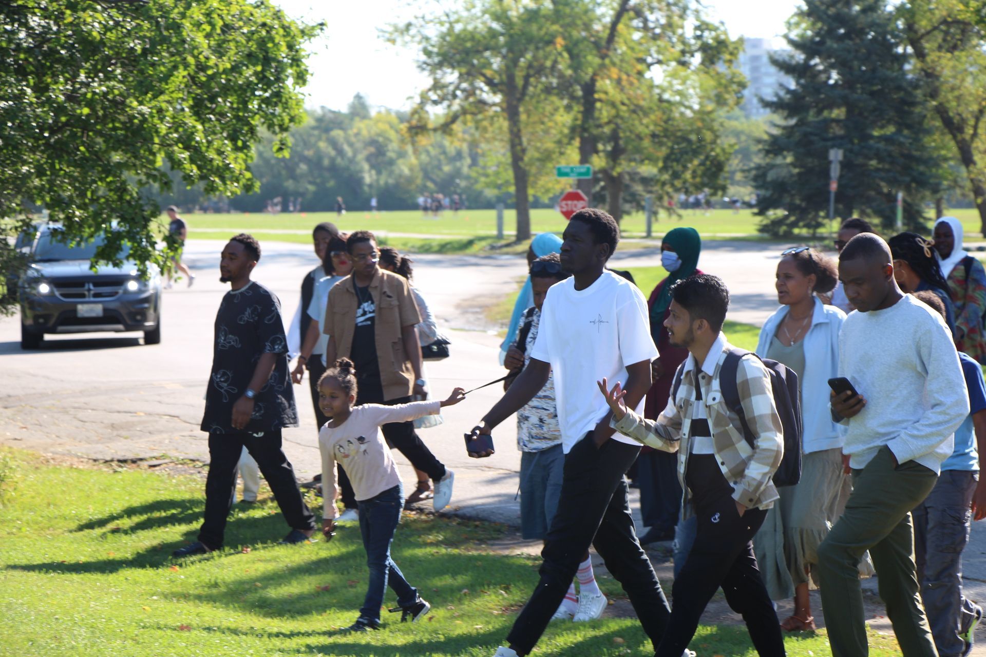 Group of people walking across a sunny park lawn, with trees and parked cars in the background.